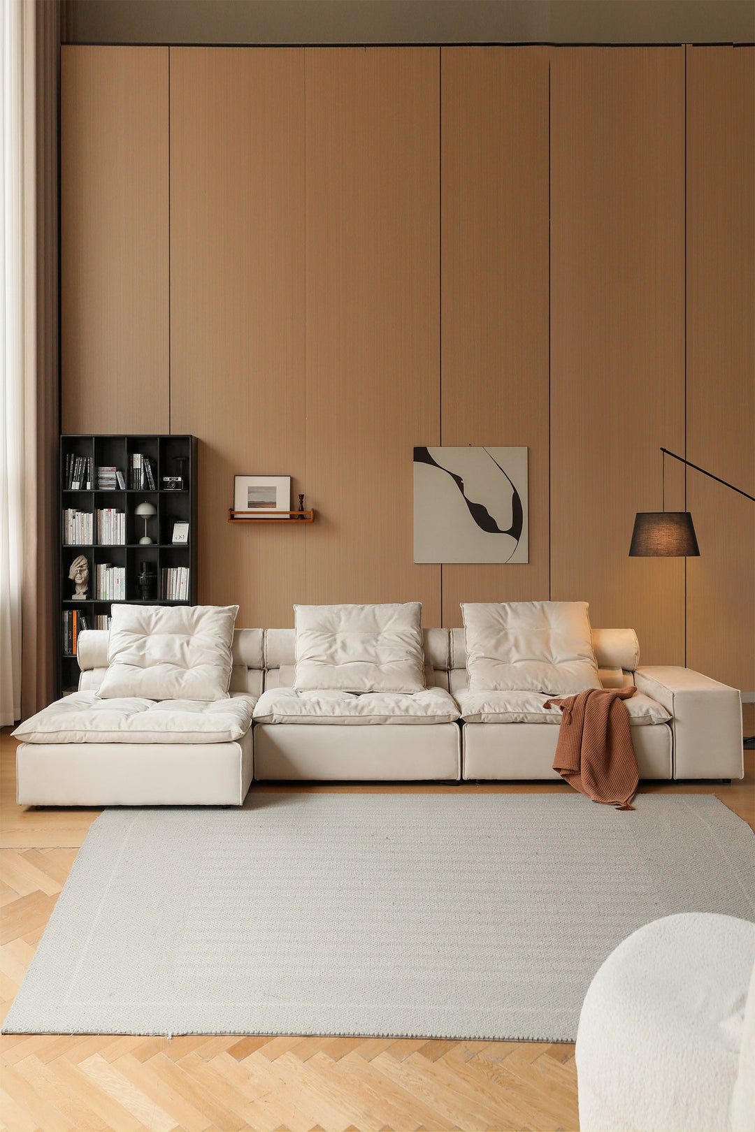 Modern living room with a beige sectional sofa, bookshelf, and artwork on a brown wall.